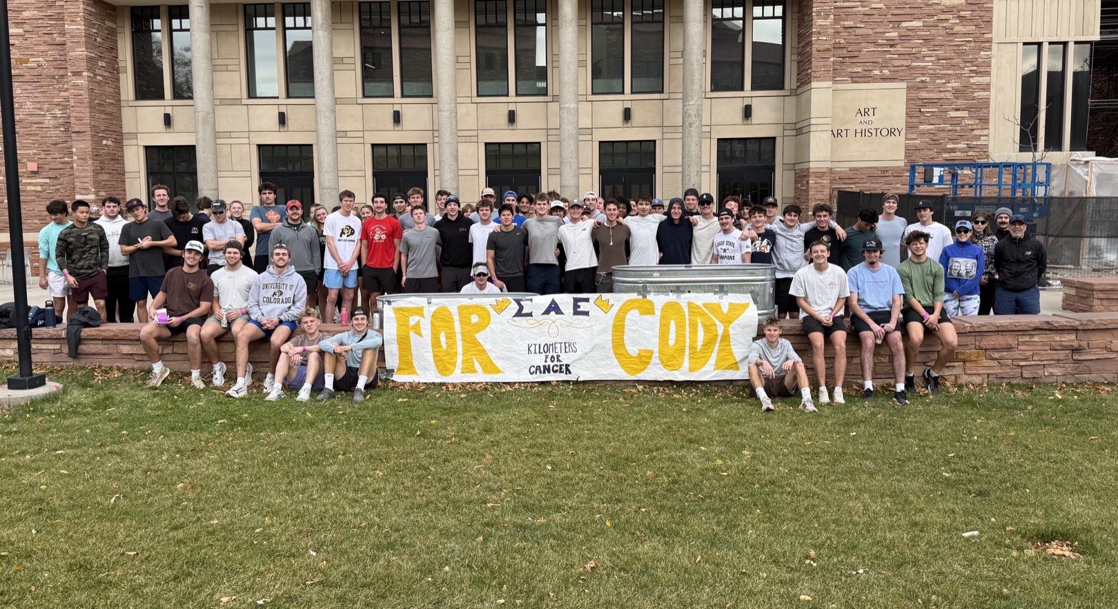 The chapter gathered outside the Art and Art History building behind a hand-painted “For Cody — Kilometers for Cancer” banner.